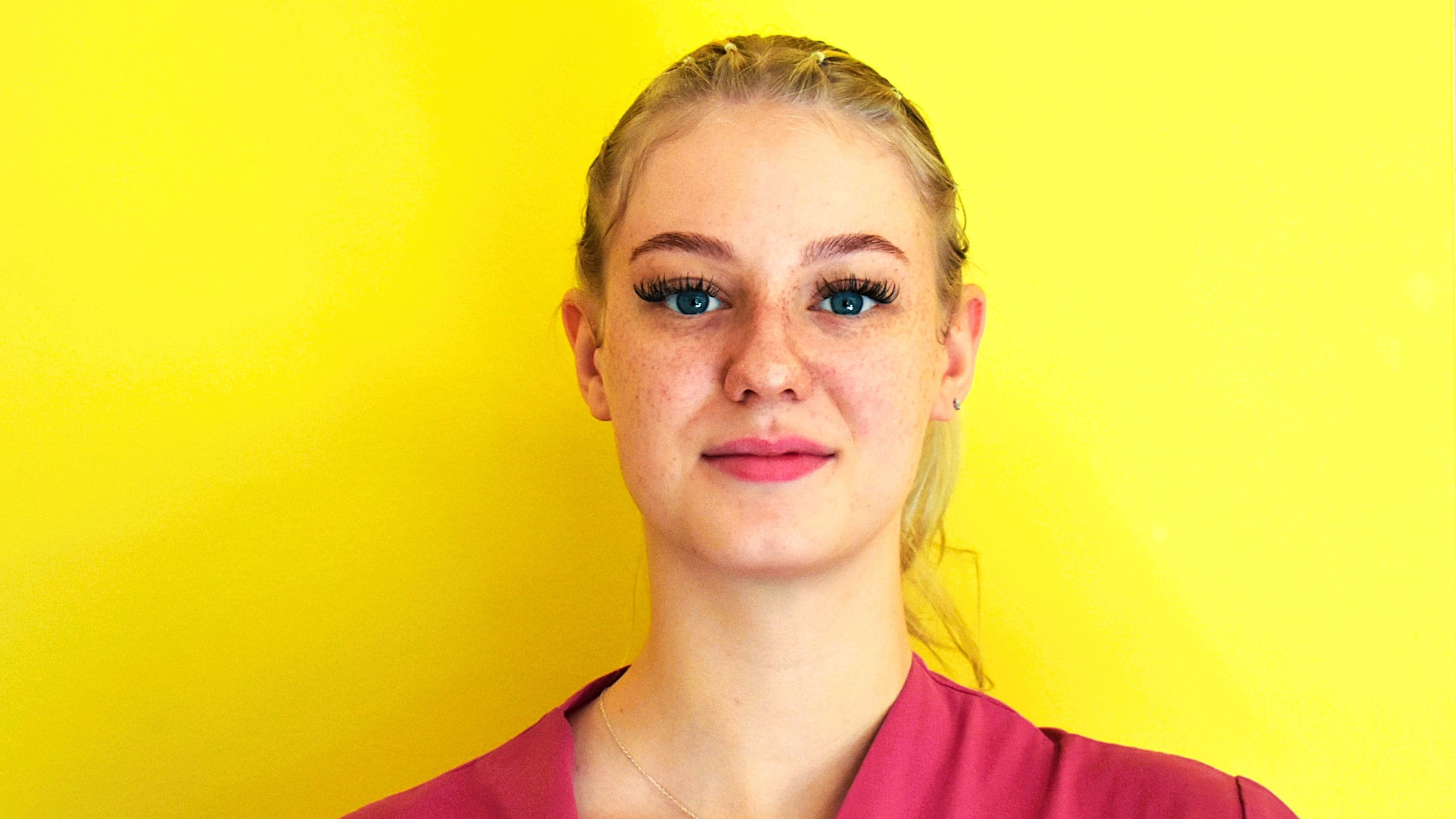Die junge Frau ist Auszubildende in unserer Senioreneinrichtung. Sie steht vor einer gelben Wand, trägt einen weinroten Arbeitskittel und lächelt in die Kamera.