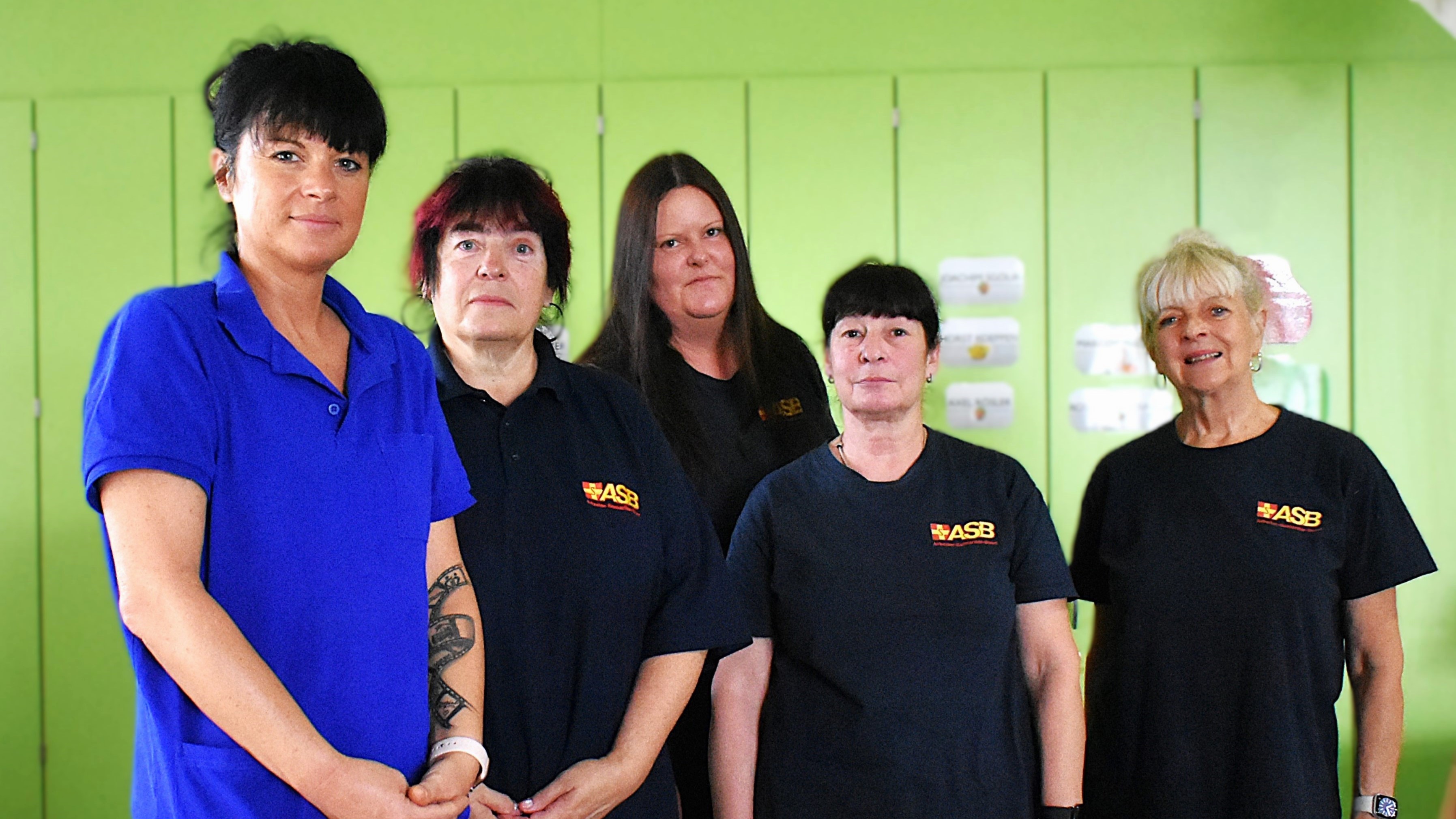 Das Team der Tagespflege Basdorfer Gärten steht in einer Reihe vor einer grünen Wand. Sie tragen blaue Arbeitskleidung. 